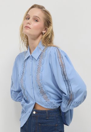 Light blue blouse with lace detailing along the front and sleeves, featuring a collared neck and button closures. Soft fabric with a loose fit.