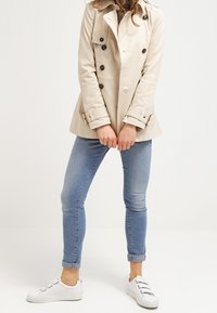 Trench-coat beige à double boutonnage, boutons noirs et bracelets aux poignets. Associé à un jean skinny bleu clair et des baskets blanches.