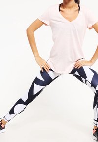 Žena v světle růžovém tričku a legínách s černo-bílým vzorem, rozkročená s nohama široko, ruce v bok.