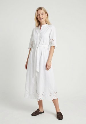 Vrouw staand, draagt een witte midi-jurk met oogjesborduurwerk, halve mouwen, een voorstrikceintuur en donkere geweven slip-on schoenen.