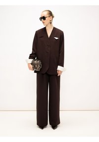 Donkerbruin op maat gemaakt pak met wijde pijpen, witte gestreepte manchetten en een decoratieve speld. Vergezeld van een metalen clutch en oversized zonnebril.