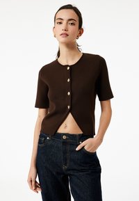 Kortærmet, brun ribbet cardigan med fem guldtonede knapper og et cropped design, parret med mørk denim højtaljede jeans.