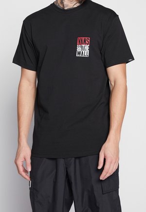 Mężczyzna w czarnej koszulce Vans z czerwono-białym logo "Vans Off The Wall" na piersi oraz w czarnych spodniach typu cargo na jednolitym tle.