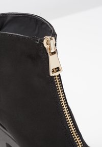 Mustat mokkajalkineet nilkat korkealla kultaisella vetskarilla sivulla, joissa on sileä pinta ja pyöreä aukko ylhäällä. Vetskarin yksityiskohta on hallitseva.