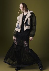 Zwarte kanten rok gelaagd over een zwart top, gecombineerd met een beige en olijfgroene schapenvachtjas. Zwarte laarzen maken de look compleet.