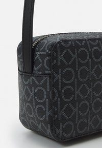 Borsa nera in materiale testurizzato con motivo a logo ripetuto in tonalità più chiara. Sottile tracolla e chiusura con cerniera.