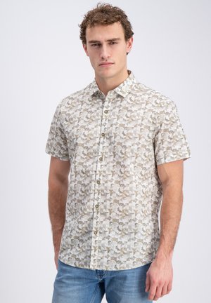 Mann trägt kurzärmliges Hemd mit grauem und beigem geometrischem Muster und blauen Jeans, steht vor einfarbig hellem Hintergrund.