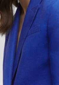 Modrý lněný blazer s klopami, jedním ramenním švem a náprsní kapsou. Texturovaná látka s hladkým povrchem. Přiléhavý střih.