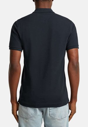 Homme portant un polo à manches courtes noir uni et un jean bleu clair, vu de dos sur un fond blanc.