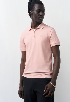 Muškarac tamne kože i dredova stoji noseći svijetloružičastu polo majicu kratkih rukava sa patentnim zatvaračem i crne hlače na jednobojnoj pozadini.