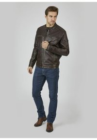Bruine leren jas met een staande kraag, rits aan de voorkant en twee borstzakken; gecombineerd met donkerblauwe jeans en bruine schoenen.
