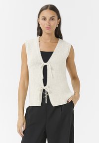 Knit sleeveless vest in cream, featuring a V-neck, center tie closure, and a relaxed fit. Textured fabric with a subtle pattern.