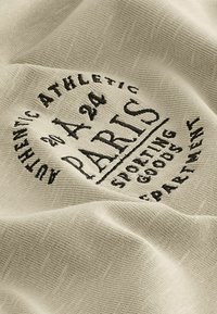 Beež kangas, millel on musta tikandi tekst. Kujunduses on sõnad "AUTHENTIC ATHLETIC", "PARIS" ja "SPORTING GOODS", tekstuurse viimistlusega.