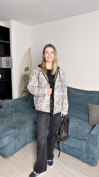 Jopica s kapuco v svetlo sivi barvi z naravnim vzorcem, kombinirana s črnimi hlačami in mornarsko modrimi supergami, drži črno torbo.