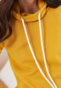 Gul hoodie med ribbat tyg, vita dragsnören och avslappnad passform, med fokus på komfort och funktionalitet.