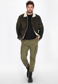 Jachetă din corduroy verde măsliniu cu guler din blană albă și nasturi negri, purtată cu pantaloni cargo și cizme negre, stând pe un fundal alb.