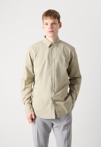 Camisa de botones de manga larga color beige claro con cuello, que presenta un logo sutil y una textura suave, combinada con pantalones grises.