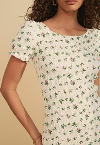 Robe florale blanche avec une texture smockée, des manches courtes bouffantes et des feuilles vertes éparpillées avec des fleurs roses.