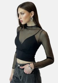 Schwarzes Mesh-Oberteil mit langen Ärmeln, körperbetontem Design, transparenter Textur und geschichtetem Ausschnitt. Kombiniert mit hochgeschnittenen schwarzen Jeans und einem silbernen Armband.