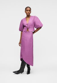 Femme portant un haut violet à manches bouffantes avec des liens et une jupe midi asymétrique assortie, accompagnés de bottes noires montantes à bout pointu.