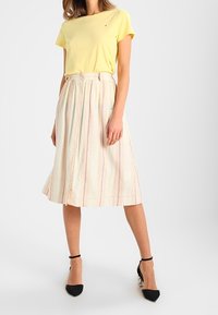T-shir coton giallo abbinato a una gonna crema con strisce verticali rosse e arancioni e chiusura frontale con bottoni. Tacchi neri a punta.
