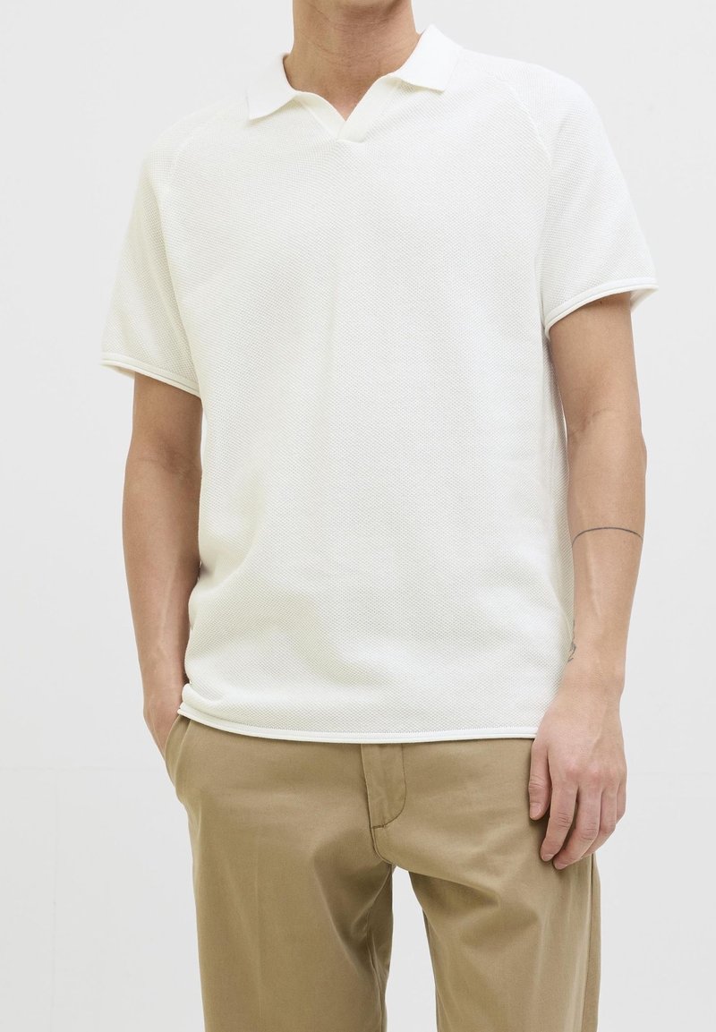 Homme portant un polo blanc texturé à manches courtes et un pantalon beige, debout, une main dans la poche, sur un fond uni.