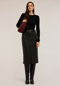 Femeie îmbrăcată într-un pulover negru cu textură în relief, fustă midi din piele neagră cu fenta frontală, cizme negre până la genunchi și ținând o geantă burgundy.