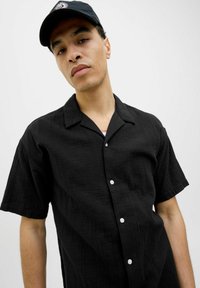 Camisa negra de manga corta hecha de una tela texturizada con un corte relajado, que presenta un collar y botones blancos en la parte delantera. Lleva una gorra negra.