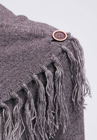 Morbid grigio tessuto lavorato a maglia con frange sciolte fissate da un grande bottone marrone su uno sfondo chiaro.