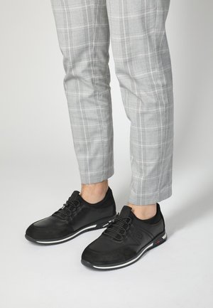 Schwarze Sportschuhe bestehen aus einer Mischung aus Leder und Wildleder, haben eine runde Spitze, eine strukturierte Oberfläche und eine kontrastierende weiße Sohle mit roten und blauen Akzenten.