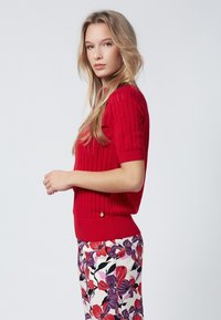 Suéter corto de manga corta en rojo con cuello redondo y un detalle dorado en el dobladillo, combinado con pantalones de flores en tonos rojos y púrpuras.