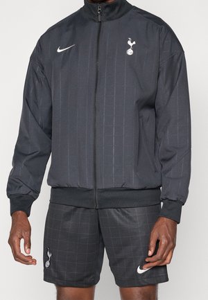 Personne portant une veste de sport noire zippée intégralement et un short assorti avec les logos blancs Nike et Tottenham Hotspur.