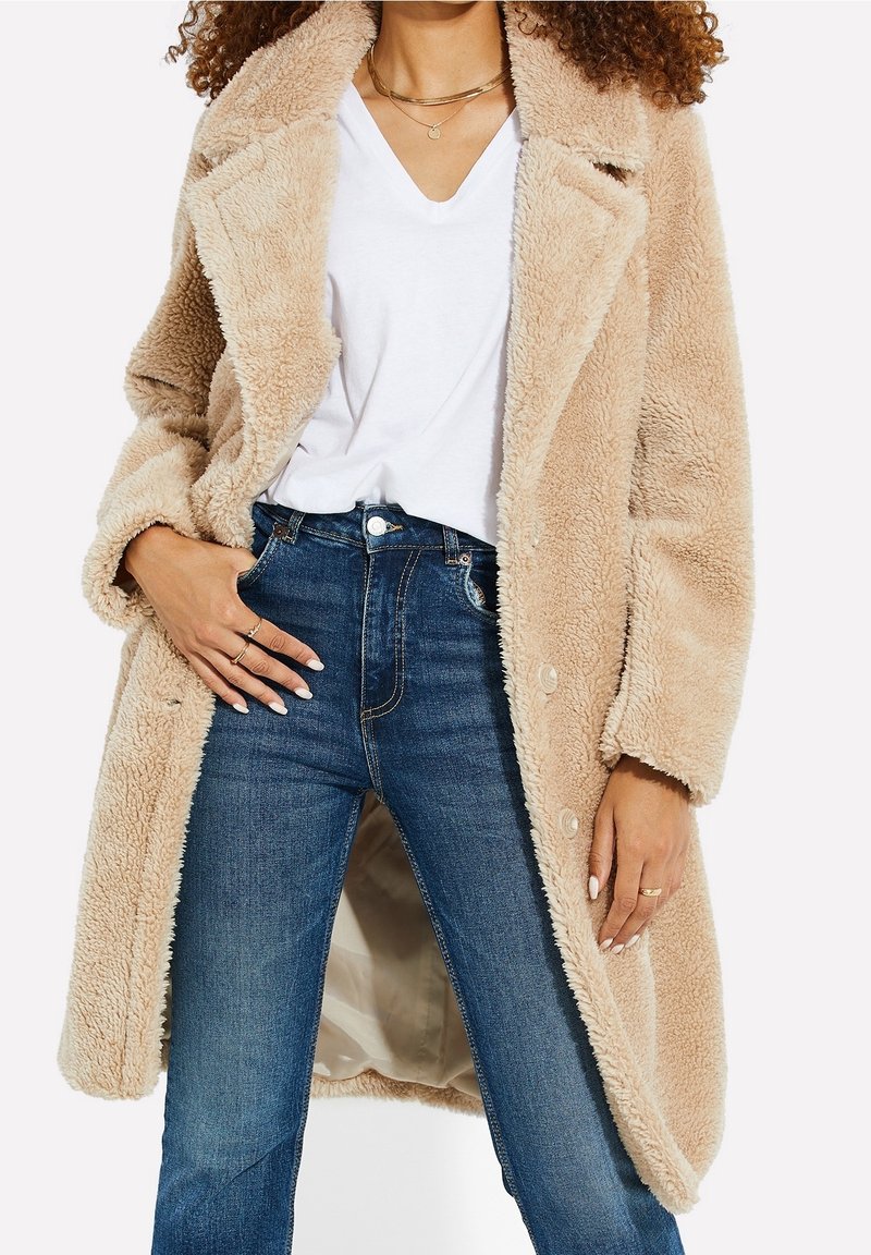 Kvinde iført beige fuzzy frakke over hvid V-hals skjorte og blå high-waisted jeans, accessorized med ringe og lagede guldhalskæder.