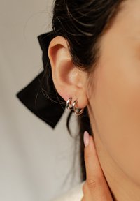 Srebrne kolczyki w kształcie obręczy o gładkim wykończeniu, dwa splatające się pierścienie, umieszczone w przekutym uchu, dopełnione czarną kokardą we włosach.