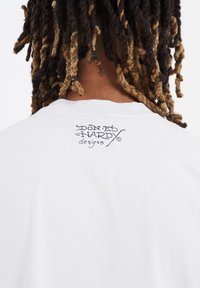 Camiseta blanca de algodón con un logo impreso en la parte posterior. El diseño incluye texto estilizado que dice "Diseños de Don Ed Hardy."
