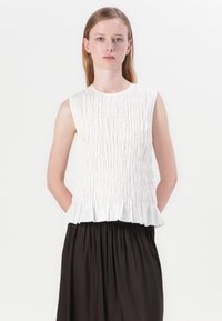 White sleeveless top featuring a smocked design with a ruffled hem, paired with a dark knee-length skirt, set against a plain background.