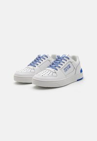 White sneakers with blue and white striped laces, perforated toe, blue accents on the heel, and text detail on the side. Flat outsole.