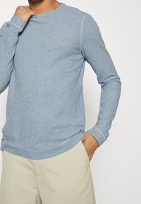 Ljusblå långärmad skjorta med texturerat tyg, rund halsringning och kontrasterande sömmar. Kombinerad med cremefärgade shorts.