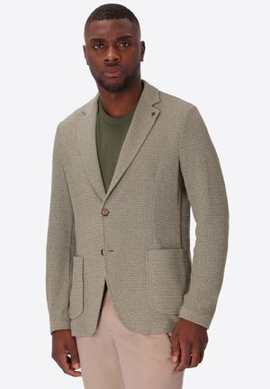 Uomo che indossa un blazer grigio chiaro strutturato, camicia verde oliva e pantaloni beige, in piedi con le mani rilassate lungo i fianchi davanti a uno sfondo semplice.