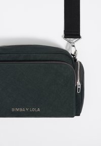 Bolso cruzado de color verde oscuro hecho de tela, que cuenta con un diseño texturizado, múltiples compartimentos con cremallera y una correa negra desmontable.