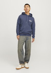Sudadera azul marino con bolsillo frontal, cordones ajustables y detalle de logo. Combinada con jeans de color azul claro y zapatillas blancas con acentos marrones.