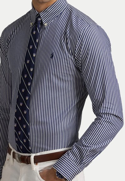 Camicia a righe blu navy e bianche con un piccolo logo, abbinata a una cravatta a motivi e una cintura, indossata con pantaloni di colore chiaro.