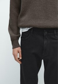 Brązowy sweter z prążkowanym wzorem noszony na białej bluzce, połączony z czarnymi dżinsami z przednią kieszenią i zapięciem na guzik.