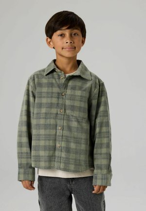 Zaļš plecu krekls ar pogām un apkakli, izgatavots no mīksta auduma, ar garām piedurknēm un brīvu piegriezumu, kombinēts ar pelēkām džinsa biksēm.