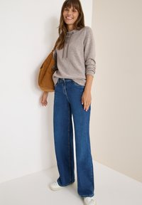 Hoodie gris clair à manches longues avec cordons, associé à un jean bleu foncé à jambes larges. Baskets blanches et sac à épaule en daim brun.