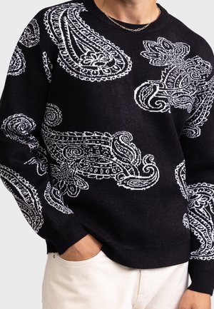 Czarny sweter z dużymi białymi wzorami paisley noszony przez osobę z ręką w kieszeni, połączony z jasnymi spodniami i złotym łańcuszkiem na szyi.