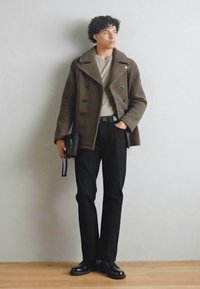 Brun dobbeltradet frakk, beige skjorte, svarte bukser, svart belte, holder en svart veske, med blanke svarte sko, mot en lys bakgrunn.