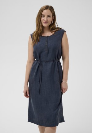 Frau trägt ein ärmelloses dunkelblaues Kleid mit Gürtel und geknöpftem Ausschnitt, steht vor einem schlichten hellgrauen Hintergrund.