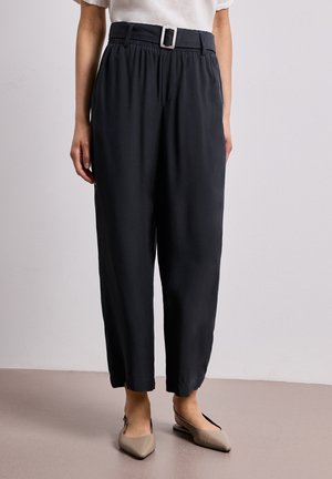 Kvinde iført sorte højtaljede taperede bukser med bælte og beige spidse slingback flade sko mod en ensfarvet baggrund.