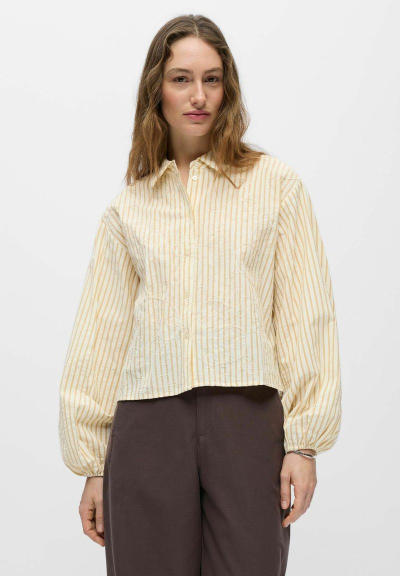 Femme portant une chemise ample à rayures beige et blanches avec des manches ballon longues et un pantalon large foncé, sur un fond uni.